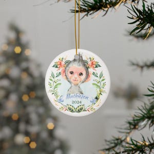 May include: A circular Christmas decoration with a watercolour illustration of a monkey surrounded by flowers. The ornament displays the name "Mathieson" and the year "2024". It is suspended by a gold-coloured string.