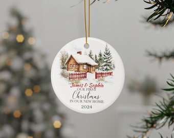 May include: A white ceramic Christmas bauble featuring a winter scene of a cabin in the woods. The bauble is inscribed with the text "James & Sophia Our First Christmas In Our New Home 2024".