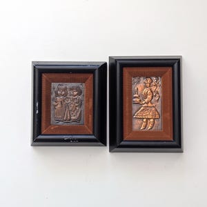 May include: Two framed copper relief art pieces. One features two figures, the other a single figure holding an object. Both are set against a brown background within black frames, creating a vintage aesthetic.