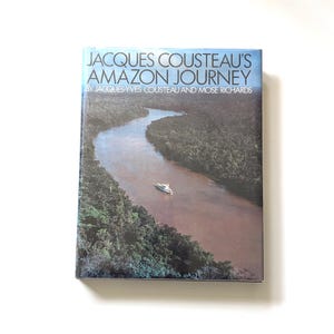 May include: A hardcover book titled "JACQUES COUSTEAU'S AMAZON JOURNEY" with a photograph of a river winding through a lush green forest. A white boat sails on the water. The book's cover has a blue and white gradient.