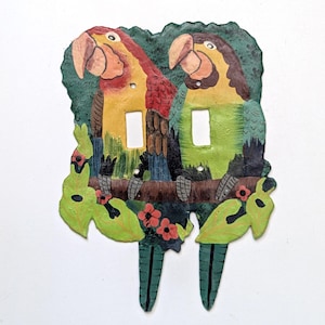 May include: A double light switch plate featuring a hand-painted design of two parrots perched on a branch. The parrots are coloured in shades of red, yellow, and green, surrounded by green leaves and small red flowers against a dark green background.
