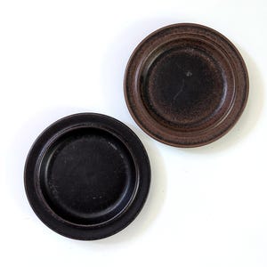 May include: Two small, round ceramic plates. One is dark brown, the other black. Both have a raised rim and textured surface. The plates are on a white background. Ideal for serving small portions.