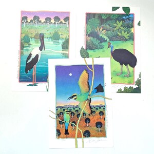 May include: Three rectangular art prints featuring Australian birds. One shows a black-necked stork in a wetland, another a cassowary in a lush forest, and the third depicts two rainbow bee-eaters in a landscape with a starry night sky.