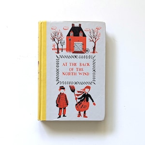 Könnte beinhalten: Ein Vintage-Hardcover-Buch mit dem Titel "At the Back of the North Wind". Das Cover zeigt ein rotes Haus mit schwarzem Dach, zwei Kinder und den Titel in einem dekorativen Rahmen. Der Buchrücken ist gelb.