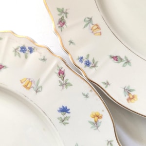 Könnte beinhalten: Nahaufnahme von zwei weißen Porzellantellern mit gewelltem Rand und Goldrand. Die Teller sind mit kleinen, farbenfrohen Blumenmustern in Gelb, Blau und Rosa verziert. Die Teller sind wahrscheinlich Teil eines Vintage-Geschirrsets.