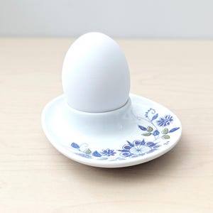 May include: A white ceramic egg cup featuring a central holder for a single egg and a wide, oval-shaped base decorated with a delicate blue and green floral pattern consisting of stylized flowers, leaves, and vines on a smooth white surface.