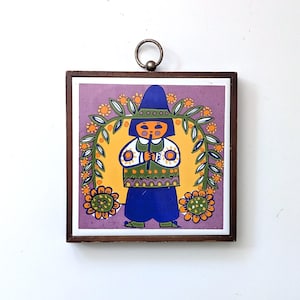 May include: A framed ceramic tile featuring a folk art illustration of a person in blue clothing and a hat, holding a flute. The background is yellow with a floral design and a purple border. The frame is brown, and it has a metal ring for hanging.