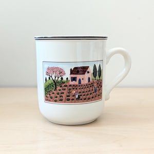 Puede incluir: Taza de cerámica blanca con borde negro y asa curva. La taza presenta una colorida ilustración de un paisaje rural con una casa, árboles y figuras. La palabra "SAPLAU" está impresa en la parte inferior de la ilustración.