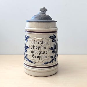 May include: A beige ceramic beer stein with a grey metal lid. The stein features a central embossed cartouche with the black text Gersten. Hopsen gibt gute Tropfen. Blue vine and hop cone motifs decorate the sides, and the lid has a textured pattern.
