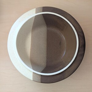 May include: Overhead view of a ceramic bowl with a white rim and a color-blocked design. The bowl features vertical stripes of cream, tan, and dark brown. The bowl is likely for serving food.