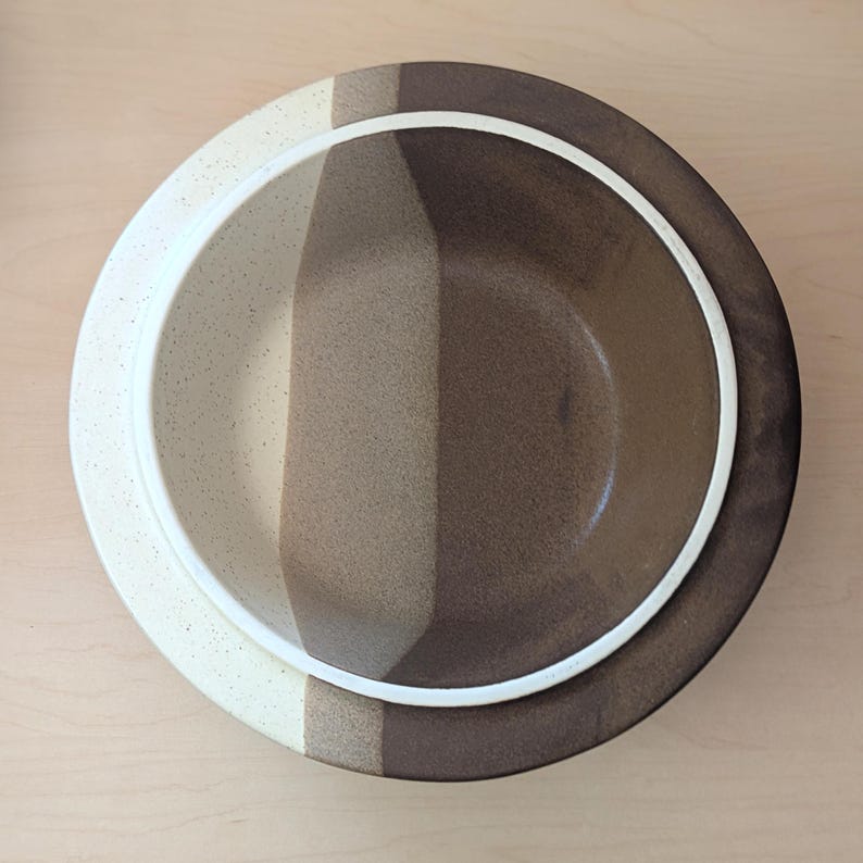 May include: Overhead view of a ceramic bowl with a white rim and a color-blocked design. The bowl features vertical stripes of cream, tan, and dark brown. The bowl is likely for serving food.