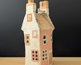Puede incluir: Una caprichosa figura de casa de cerámica con una fachada blanca y rosa, con múltiples ventanas y un tejado texturizado. La casa tiene dos chimeneas con detalles amarillos y un pequeño pájaro posado en el tejado. Es un objeto decorativo.