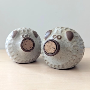 Puede incluir: Dos tapones de botella de cerámica con forma redonda y un inserto de corcho. Los tapones son de color gris claro con detalles marrones y una superficie texturizada. Los tapones tienen un diseño caprichoso, que se asemeja a un animal pequeño.