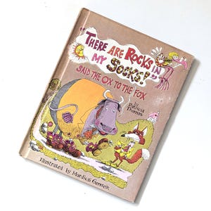 Puede incluir: Un libro infantil vintage titulado "There Are Rocks in My Socks?" con ilustraciones de un buey, un zorro y otros personajes. La portada es de color canela con ilustraciones y texto coloridos.
