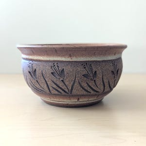 May include: A brown ceramic bowl with a textured surface and a floral design carved around the middle. The bowl has a wide rim and a slightly flared shape. The color is a mix of brown and tan.