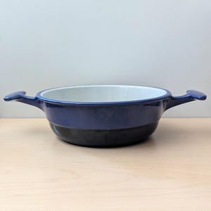 Può includere: Una pirofila in smalto blu e nero con due manici. L'interno è bianco e l'esterno è diviso in una sezione superiore blu e una sezione inferiore nera. La pirofila è su una superficie di legno chiaro.