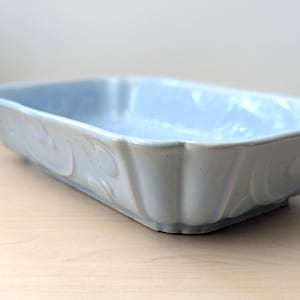 Op de afbeelding: Een lichtblauwe rechthoekige keramische ovenschaal. De schaal heeft een geschulpte rand en een decoratief krulpatroon aan de zijkanten. De lege schaal staat op een lichtgekleurd houten oppervlak.