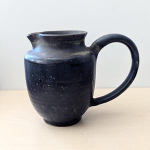 May include: A matte black ceramic pitcher with a rounded body, a curved handle, and a small pouring spout. The surface features a textured, dark finish with subtle variations in tone and light speckling around the rim and upper body.