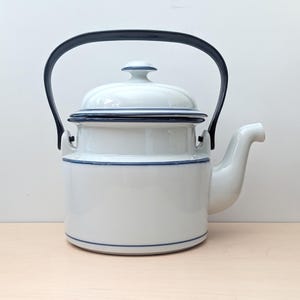 May include: White ceramic teapot with a black handle and lid. The teapot has a rounded body with a spout and is accented with blue trim. The handle is arched, and the lid has a small knob. The teapot is designed for brewing tea.