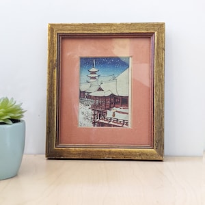 May include: Framed Japanese woodblock print depicting a snow-covered temple scene. The artwork features a pagoda and buildings with red accents, set against a blue sky with falling snow. The frame is gold-colored.