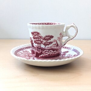 Puede incluir: Un juego de taza y platillo blancos con un diseño de paisaje burdeos. La taza tiene un asa en forma de lazo y un borde festoneado. El platillo también tiene un borde festoneado. El diseño presenta árboles, un puente y una casa.