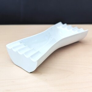 May include: White ceramic ashtray with a wavy design and multiple indentations for cigarettes.