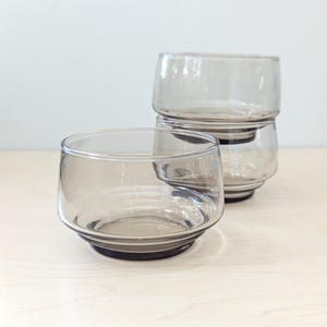 Könnte beinhalten: Drei klare, grau getönte Glasbecher. Ein Becher befindet sich im Vordergrund, mit zwei dahinter gestapelten. Die Gläser haben eine abgerundete Form mit einem dicken, dunkelgrauen Boden. Der Hintergrund ist hell und neutral.