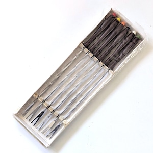 May include: A boxed set of fondue forks. The forks have long, stainless steel tines and dark brown handles. The handle ends are capped with various colours, including red, yellow, and green. The box is made of cardboard.