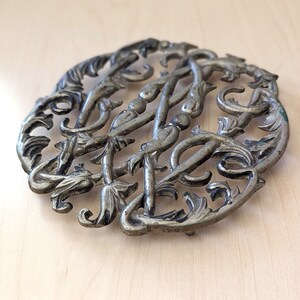 May include: Silver-toned metal trivet with an intricate floral design. The trivet is round and has a delicate, ornate pattern.