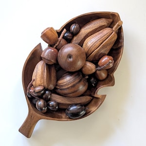 May include: A wooden leaf-shaped bowl filled with various wooden fruits and nuts. The bowl and the wooden objects are in shades of brown. The arrangement includes what appear to be bananas, apples, and other unique shapes.