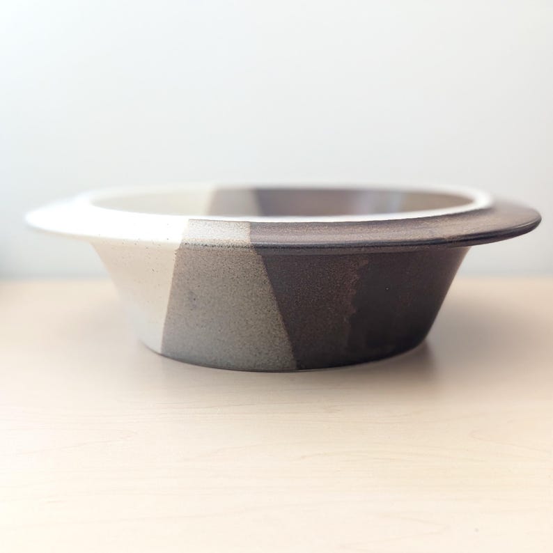 May include: A ceramic bowl with a wide, flat rim and a geometric design. The bowl is divided into three sections of color: white, light gray, and dark brown. The bowl is sitting on a light-colored wooden surface.
