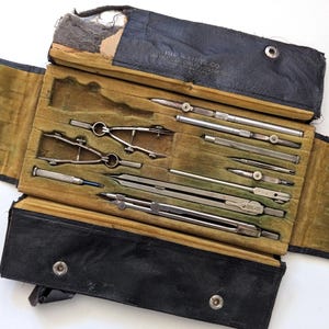 May include: A vintage drafting instrument set housed in a black leather-like folding case with a mustard-colored velvet interior. The set includes various metal compasses, dividers, and precision tools, with the text THE A. LIETZ CO. visible.