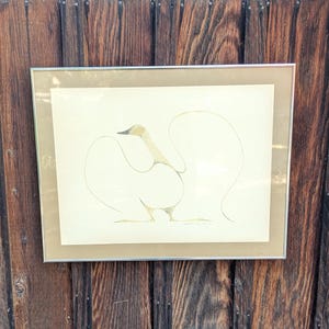 May include: Framed artwork featuring a minimalist illustration of a swan. The swan is depicted with soft, flowing lines in shades of beige and gold against a cream background. The artwork is encased in a silver-toned frame.