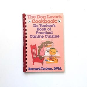 May include: A spiral-bound cookbook titled The Dog Lover's Cookbook: Dr. Tonken's Book of Practical Canine Cuisine by Bernard Tonken, DVM. The cover features a pink background with an illustration of a dog wearing an apron cooking at a table.