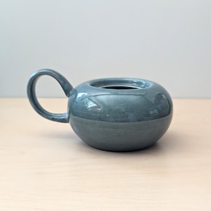 Könnte beinhalten: Eine teal-farbene Keramik-Teekanne mit rundem Körper und gebogenem Griff. Die Teekanne hat eine breite Öffnung oben und steht auf einer hellen Holzoberfläche. Die Teekanne ist ein dekorativer Gegenstand.