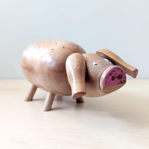 Puede incluir: Un alfiletero de madera en forma de cerdo con el hocico pintado de rosa y los ojos pintados de blanco. El cerdo tiene cuatro patas y está de pie sobre una superficie marrón claro.