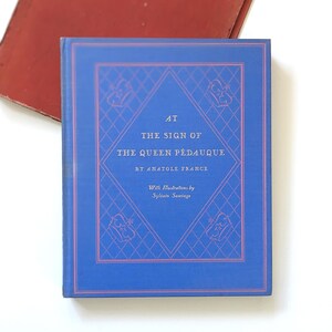May include: A hardcover book titled "At The Sign of The Queen Pédauque" by Anatole France. The book has a blue cover with a diamond-shaped design and text in white. The book is partially visible, with a red surface in the background.