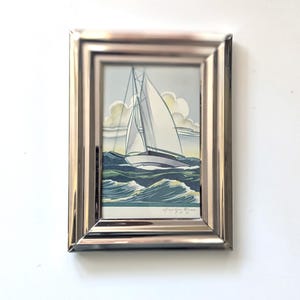 May include: A framed artwork featuring a sailboat on a wavy sea under a cloudy sky. The sailboat has white sails and is set against a light blue background. The frame is silver-colored.