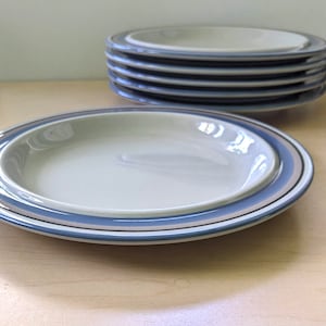 Może przedstawiać: Zestaw sześciu białych ceramicznych talerzy z niebieskimi i szarymi paskami wokół krawędzi. Talerze są ułożone jeden na drugim.