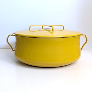 May include: A bright yellow enamel pot with a matching lid. The pot has two side handles and a wire handle on the lid. The pot is round and sits on a white surface. The pot is a vibrant yellow color.