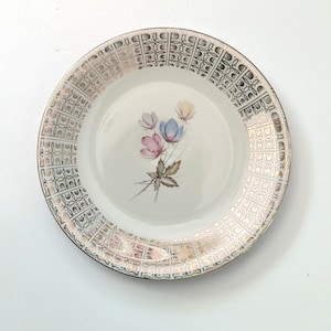Op de afbeelding: Een vintage witte bord met een gouden en zilveren geometrische rand. Het midden heeft een bloemenontwerp met roze, blauwe en gele bloemen. Een klassiek stuk om te serveren of tentoon te stellen.
