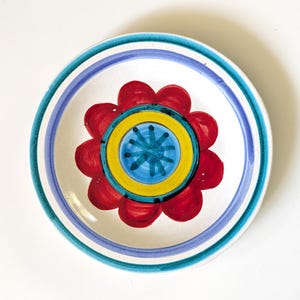 May include: A white ceramic plate with a hand-painted floral design. The plate features a red flower with yellow and blue accents in the center, surrounded by blue and teal stripes along the rim. The plate is round.