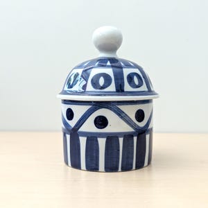 May include: A white ceramic lidded jar featuring a hand-painted dark blue geometric pattern. The design includes vertical stripes at the base, a series of triangles with central dots in the middle, and a dome-shaped lid with a round knob handle.