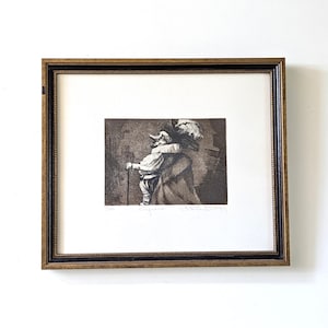 Pode incluir: Uma gravura em preto e branco emoldurada retratando uma figura em traje histórico com um grande colarinho, um chapéu com penas e uma bengala. A obra de arte é apresentada numa moldura retangular dourada e preta com paspatur branco.