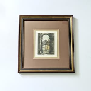 May include: A framed black and white print of an architectural scene. The print shows a view through an archway, with a building and a street visible in the distance. The print is matted and framed in a dark wood frame.