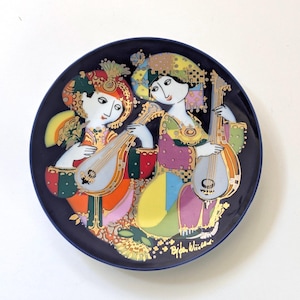 May include: Decorative plate with a dark blue rim featuring two figures playing stringed instruments. The figures are dressed in colorful attire with gold accents. The plate has a white background and the artist's signature is visible.