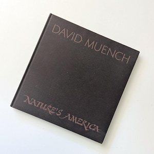 May include: A dark brown hardcover book with the title "DAVID MUENCH NATURE'S AMERICA" in a lighter brown font. The author's name is above the title. The book is angled, set against a white background.
