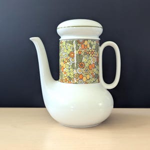 Puede incluir: Cafetera de cerámica blanca con un estampado floral en amarillo, naranja y verde. La cafetera tiene un pico curvo, un asa y una tapa con ribete dorado. El diseño floral envuelve la parte media de la cafetera.