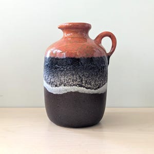 Puede incluir: Un jarrón de cerámica con asa, que presenta un degradado de color. La parte superior es naranja, pasando a negro, luego a blanco y terminando con una base marrón oscuro. El jarrón tiene un cuerpo redondeado y un asa pequeña y curvada.