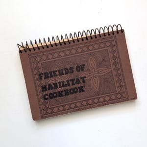 May include: A brown spiral-bound cookbook with black wire binding. The cover features a black geometric border design and the text "FRIENDS OF HABILITAT COOKBOOK" printed in a bold, blocky font. The cover has a matte texture and rectangular shape.
